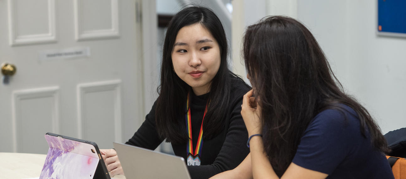 Two students sitting at a desk with laptops, chatting to each other. 