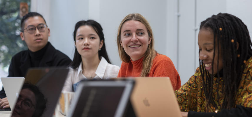 Four people sitting on one side of a meeting table, all at laptops. Three are looking to the right and smiling. In the foreground on the right, another smiles and looks at her notes.