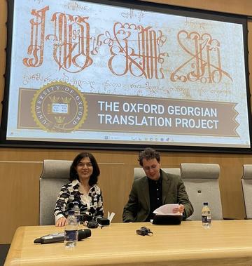 Lia Chokoshvili and Oliver Matthews sitting at a large wooden desk. Lia is smling at the camera and Oliver is looking down at a book he has opened, smiling.