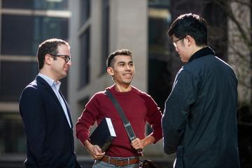 Two students and a teacher in a group, standing and chatting outside department buildings. 