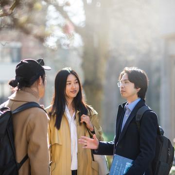 Three students standing outside under a large tree. They are wearing light winter coats and carrying backpacks.