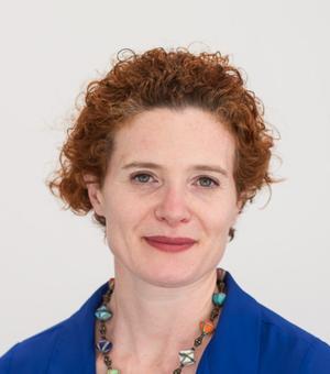 Anna Lora-Wainwright has curly auburn hair that she has tied back. She is wearing a royal blue shirt with a necklace made from various multicoloured beads. 