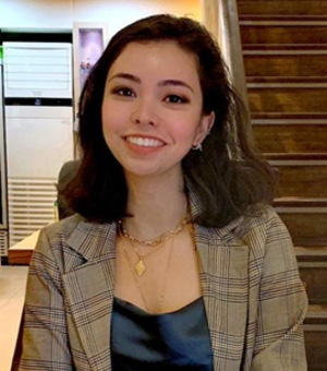 Eliza has shoulder length brown hair and brown eyes. She is wearing a dark blue silk shirt, brown checked blazer and gold jewellery. She is smiling at the camera. 